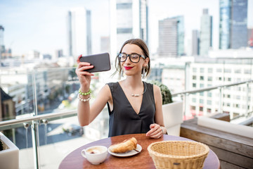 Happy businesswoman making selfie portrait sitting on the terrace during the breakfast with skyscrapers on the background in Frankfurt
