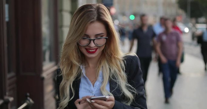 Attractive blonde woman with red lipstick, in a business suit walking down the crowded central street, passing by the street musician and using her phone, reacting happily to the message.