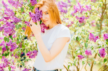Fototapeta premium Portrait of young attractive cute redhair girl outdoor in park in blooming lilac bushes in summer sunny day. Resting among paradise flowers. Fragrance and fresh smell. Tenderness, innocence, airness.