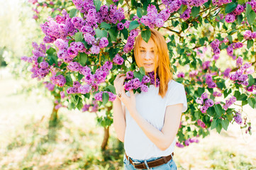 Obraz na płótnie Canvas Portrait of young attractive cute redhair girl outdoor in park in blooming lilac bushes in summer sunny day. Resting among paradise flowers. Fragrance and fresh smell. Tenderness, innocence, airness.