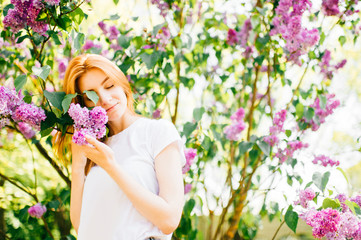 Beautiful young tender cute redhead girl with eyes closed in park in blooming lilac bushes in sunny day on weekend. Relaxing in paradise flowers. Fragrance in air. Tenderness, innocence, airness.