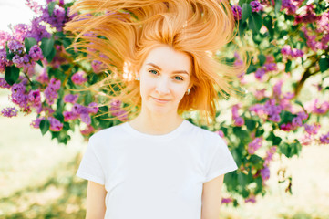 Fototapeta premium Young beautiful tender redhead cute girl getting fun. Playing with hair, looking at camera in park outdoor in summer sunny day on weekend with lilac blooming tree on bright background.