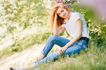Naklejka na ściany i meble Beautiful and attractive foxy cute redhead girl in casual clothes sitting and looking at camera in park outdoor under lilac bushes in summer sunny day. Blue jeans with belt. Youth fashion style. .