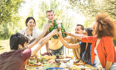 Young multiracial friends toasting at barbecue garden party - Friendship concept with happy people having fun at backyard bbq summer camp - Food and drinks fancy picnic lunch - Focus on beer bottles
