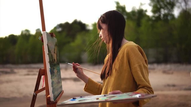 A young artist draws a palette knife still life, a girl creates a work of art near the sea or river. Person placed Malbert in the open air in order to draw from nature
