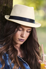 Fototapeta premium Beautiful young woman with a hat sitting in a park, holding a bottle with her favorite juice and relaxing