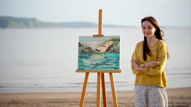 Young woman artist and her canvas painted en plein air against bright sunlight, water and green nature