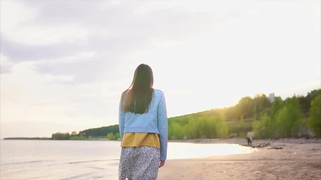 A thin teenager with long hair in modern clothes strolls along a deserted shoreline with wet sand, the girl slowly walks along the beach near the sea or the river