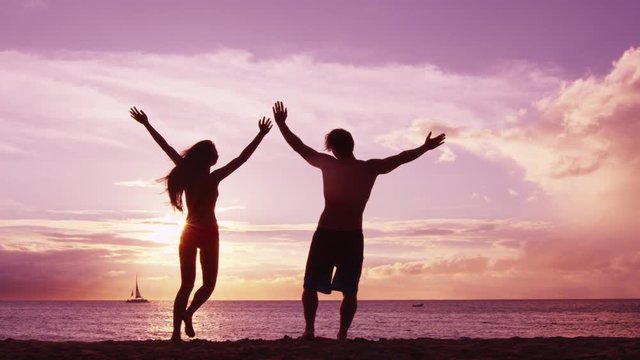 Happy couple jumping on beach enjoying sunset on summer travel vacation holiday. Happiness and bliss concept video with people jumping of joy celebrating in silhouette. SLOW MOTION RED EPIC.