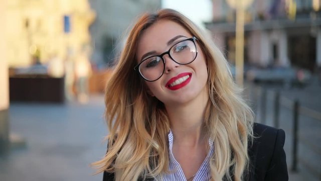 Close up view of a beautiful nerdy like attractive blonde woman in glasses with red lips looking right to the camera and smiling charmingly. Positive mood, being happy.