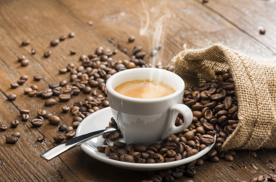 steaming cup of coffee with burlap sack and coffee beans on wooden table.