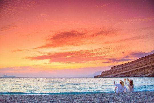 Two women taking photos of the amazing sunset at the beach of Matala, Crete, Greece.