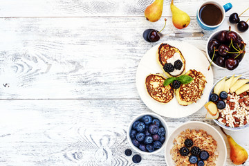Healthy summer breakfast with copy space. Top view of oatmeal, puffed rice cereal, pancakes with blueberry, cherry and pear, cup of black coffee on white wooden table.