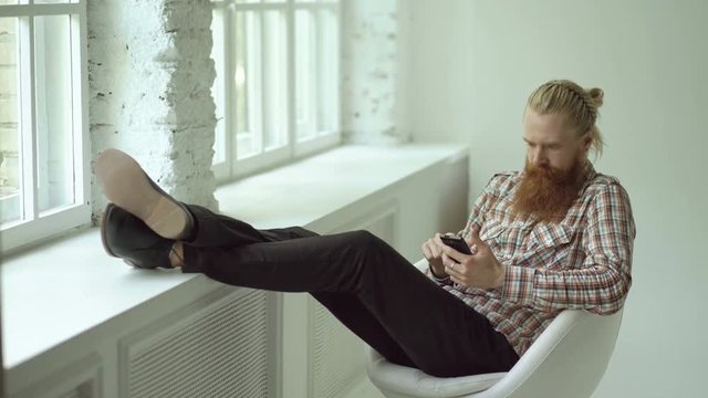 Bearded young hipster man sms texting using smartphone while sitting in offce chair with his legs on windowsill