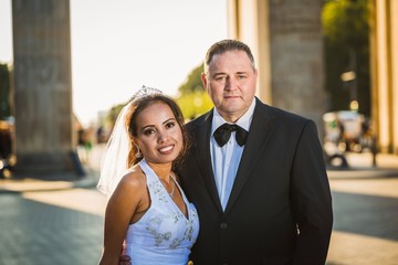 Portrait of a happy multicultural wedding couple at Brandenburger Gate with back sun light. Newlyweds. The two married are looking at the camera. © arianarama