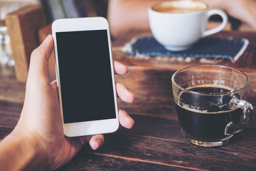 Mockup image of hand holding white mobile phone with blank black screen with coffee cup on wooden table in vintage cafe