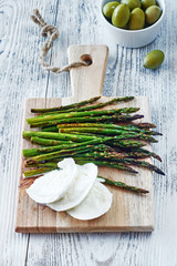 Baked asparagus with mozzarella on wooden board and green olives on white wooden table. Vertical closeup of healthy appetizer. 