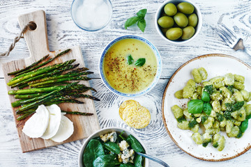 Top view of healthy green lunch or dinner concept. Gnocchi with pesto, spinach salad with blue cheese, baked asparagus with mozzarella, green puree soup and olives on white table. 