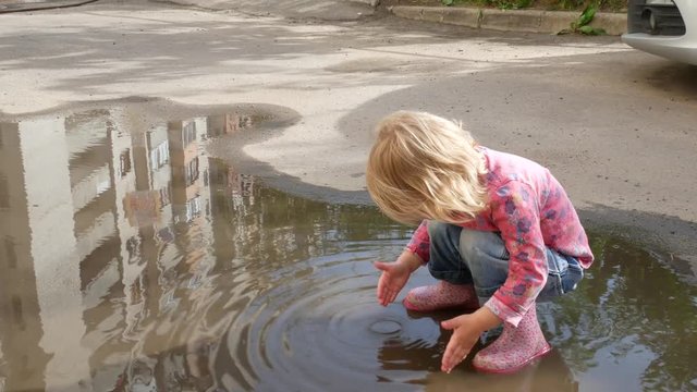 The child washes his hands in a puddle. The girl splashes in a puddle with her hands.
