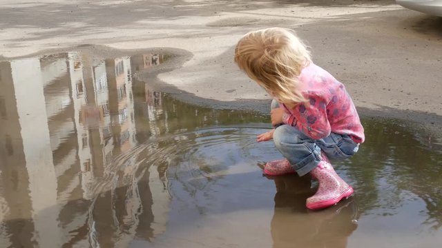 The child washes his hands in a puddle. The girl splashes in a puddle with her hands.