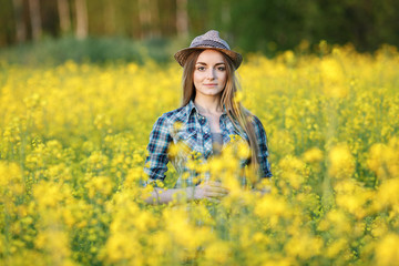 Fototapeta premium Attractive young blonde woman in blue plaid shirt straw hat enjoying her time on bright colorful blossoming yellow green meadow hard noon sun.