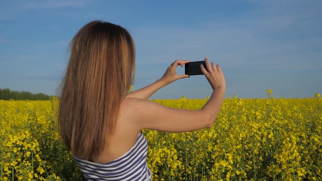 A young beautiful girl stands in a blooming field and takes photos on the phone.