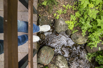 Girl in white slippers and jeans standing on a wooden bridge, which is laid through a small forest stream in the park. Top view.