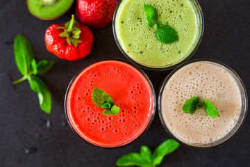 Close-up three glasses with smoothies, banana, kiwi, strawberries, on a black stone surface, decorated with mint, next to lie fresh berries and fruits, a tasty bright dessert, healthy food, restaurant