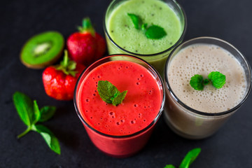 Close-up three glasses with smoothies, banana, kiwi, strawberries, on a black stone surface, decorated with mint, next to lie fresh berries and fruits, a tasty bright dessert, healthy food, restaurant