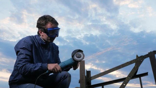A young man polishes a metal angle grinder at sunset. Beautiful sky behind the worker.