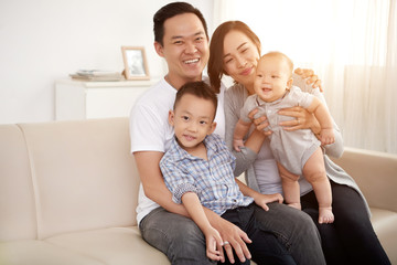 Portrait of happy Asian family with two children posing on sofa at home looking at camera in sunlight