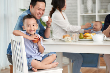 Obraz na płótnie Canvas Portrait of happy Asian boy smiling to camera, holding apple while sitting at breakfast table with family