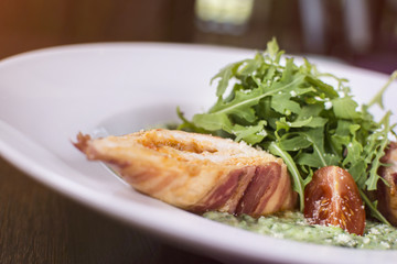 Roasted chicken breast wrapped in bacon slices with greens, tomatoes, and sauce. Beautifully served meal at the restaurant.