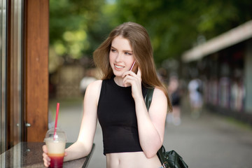 Young beautiful stylishly dressed girl is walking along the streets of the city, drinking juice, coffee, talking on mobile phone