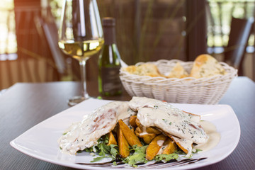 Grilled chicken breast covered with sauce and potatoes in a plate on wooden table. Beautifully served meal at the restaurant. Bread and wine in the background.