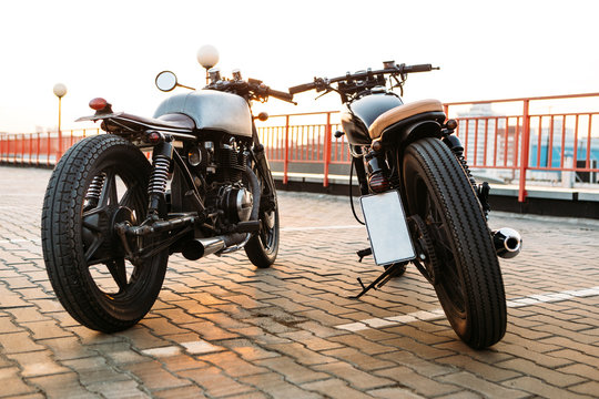 Back view of two vintage custom motorbike cafe racer motorcycle looking in same direction on empty rooftop parking lot with backlight sun during sunset. New urban style. Hipster lifestyle.