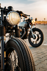 Vintage custom motorcycle caferacer motorbike with lamp lights turned on. One with grill headlight another with tape cross over optic on empty rooftop parking lot during sunset. Hipster lifestyle.