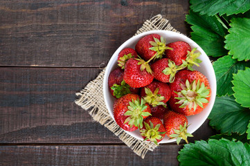 A bowl of ripe strawberry among the green leaves on a dark wooden table. Top view.