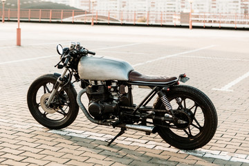 Obraz na płótnie Canvas Silver vintage custom motorcycle motorbike cafe racer on empty rooftop parking lot at city center surrounded by urban environments midtown buildings. Hipster style, student dream, wild lifestyle.