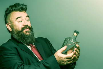 Adult bearded man in suit looking at a bottle of alcohol in his hand on a green background