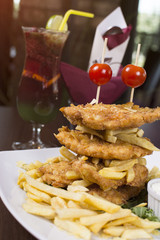 Fried pork chop in breadcrumbs, served with French fries. Stabbed with sticks on a white plate and sour cream. Beautifully served meal at the restaurant. Coctail in background.