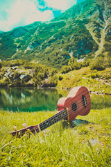 Romantic view of the Ukulele guitar at the mountain nature green meadow. Photo depicts musical instrument Ukulele small guitar at the amazing pure mountain lake Shore and the blue sky background.