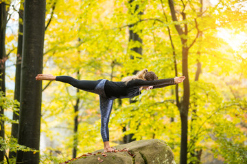 Yoga outdoors - sporty fit female doing asana Virabhadrasana 3 in autumn park on the big boulder
