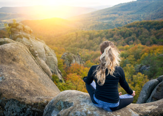 Naklejka na ściany i meble Young fit girl is practicing yoga and doing asana Siddhasana on the top of the high rocky mountain in the evening. Beautiful sunset, autumn forests, rocks and hills on the background