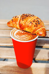 Red paper cup with coffee and chocolate croissant. Coffee to go. Tasty hot beverage on wooden table...