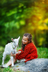 The girl sits on a large gray stone in the middle of the forest and offers to eat grass to a small white goatling