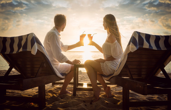 Paar mit Cocktails am Strand auf Strandliegen schaut den Sonnenuntergang im Urlaub an