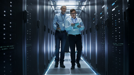 In Data Center Two IT Engineers Walking Through Rows of Server Racks. They Work on Tablet Computer...