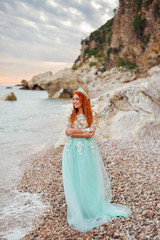 Fototapeta na wymiar Young beautiful red-haired woman in a luxurious dress standing on a rocky shore of the Adriatic Sea