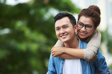 Obraz premium Portrait of young man carrying pretty Asian woman on back, giving piggyback ride on date, having fun